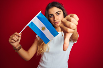 Young beautiful woman holding argentine flag over red isolated background with angry face, negative sign showing dislike with thumbs down, rejection concept