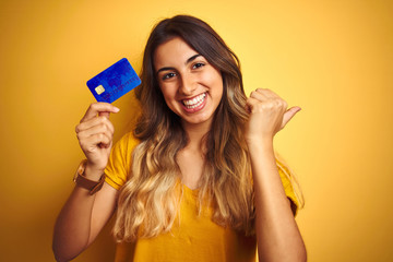 Young beautiful woman holding credit card over yellow isolated background pointing and showing with thumb up to the side with happy face smiling
