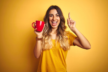 Young beautiful woman holding  red cup of coffee over yellow isolated background surprised with an idea or question pointing finger with happy face, number one