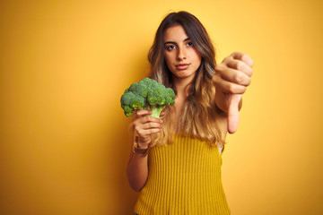 Young beautiful woman eating broccoli over yellow isolated background with angry face, negative...