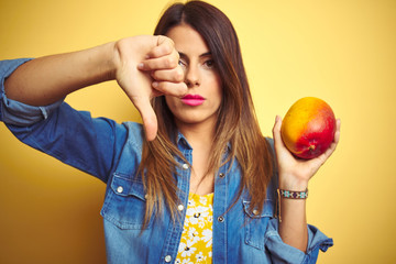 Young beautiful woman eating fresh healthy mango over yellow background with angry face, negative...