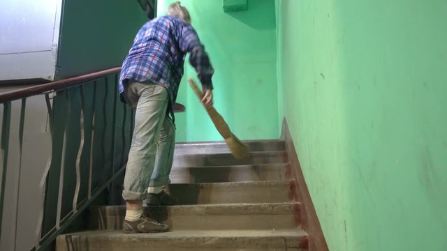 A young woman in blue jeans and a cage shirt swings and washes the floor on a staircase next to an elevator in a Stalinist-built apartment building, Cleaning public spaces by a labor immigrant cleane