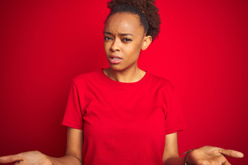 Young beautiful african american woman with afro hair over isolated red background clueless and confused with open arms, no idea concept.