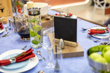 a table with a blue tablecloth is festively served, steel appliances and a salt shaker, a white and bare plate with a red napkin, a decanter filled with lime water and a black plate for copyspace.
