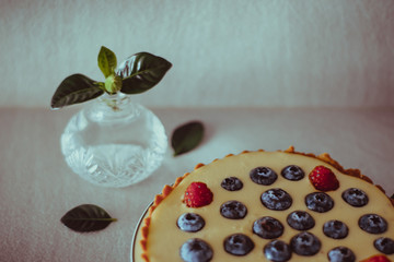 Homemade berry cake with raspberries and blueberries and crystal vase with small bud of gardenia on gray minimalistic background