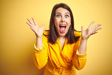 Young beautiful brunette woman wearing elegant dress over yellow isolated background celebrating crazy and amazed for success with arms raised and open eyes screaming excited. Winner concept