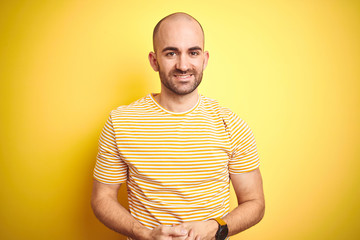 Young bald man with beard wearing casual striped t-shirt over yellow isolated background with hands together and crossed fingers smiling relaxed and cheerful. Success and optimistic