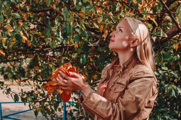 Blonde woman wearing a brown leather jacket smiling and holding fallen leaves in her hands. Autumn...