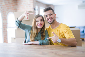 Young couple sitting on the table movinto to new home with carboard boxes behind them smiling making frame with hands and fingers with happy face. Creativity and photography concept.