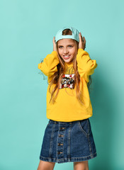 Funny teenage girl in a baseball cap and yellow hoodie covered her ears with her hands on a blue background.