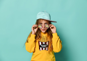 Funny teenager girl in a baseball cap, funny trendy casual glasses and yellow hoodie posing on a blue background. 