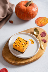 Portion of delicious bright orange pumpkin cheesecake on plate cut from whole cake, decorated with chocolate on white background with pumpkin and autumn leaves