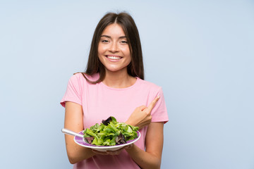 Young woman with salad over isolated blue background pointing to the side to present a product