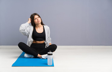Young sport woman sitting on the floor with mat having doubts and with confuse face expression