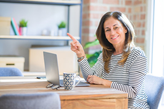 Middle age senior woman sitting at the table at home working using computer laptop very happy pointing with hand and finger to the side