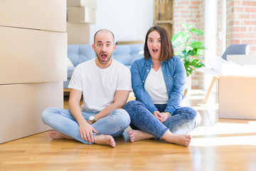 Young couple sitting on the floor arround cardboard boxes moving to a new house afraid and shocked with surprise expression, fear and excited face.