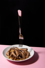 Pile of oily fish on white plate on pink tabletop with black background. Floating fork above. Abstract conception.