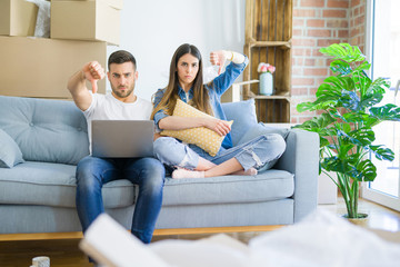 Young beautiful couple relaxing sitting on the sofa of new house using computer laptop with angry face, negative sign showing dislike with thumbs down, rejection concept