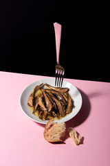 Pile of oily fish on white plate on pink tabletop with black background. Floating fork above. Abstract conception.