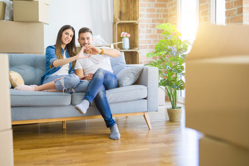 Fototapeta premium Young beautiful couple relaxing sitting on the sofa around boxes from moving to new house smiling in love showing heart symbol and shape with hands. Romantic concept.