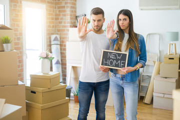 Fototapeta na wymiar Young beautiful couple holding blackboard with new home text at new house with open hand doing stop sign with serious and confident expression, defense gesture