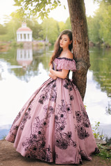 Fototapeta premium A beautiful young woman in a 19th-century dress stands with a book in a park. Romantic girl at the pond.