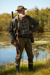 Senior man with gun going to hunt on wild ducks on lake. Male looking away. Forest lake background