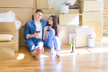 Fototapeta na wymiar Young couple sitting on the floor of new house around cardboard boxes, celebrating drinking a glass of wine and smiling happy for new apartment