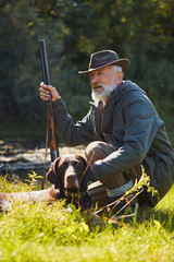 Senior good-looking hunter with his tracker dog in hunt on wild ducks. Sitting near lake, wearing...