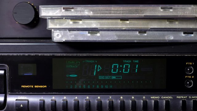 Close POV shot of a remote control activating a CD player, with track 1 starting to play on the display, and CD cases in front of the amplifier volume control.