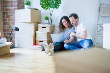 Middle age senior romantic couple in love sitting on the apartment floor with boxes around and using computer laptop smiling happy for moving to a new home