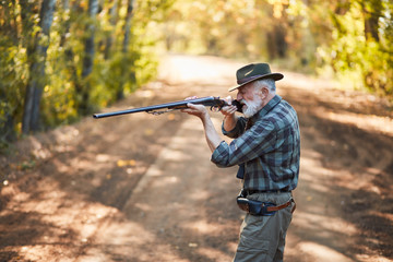 Side view on senior hunter man aims at trophy bird in autumn sunny forest, pointing gun at bird.