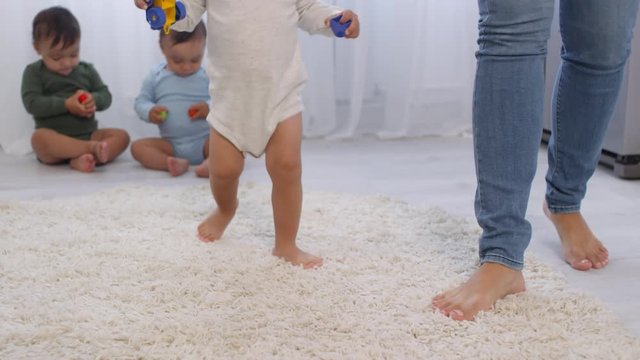 Close-up legs shot of unrecognizable barefoot toddler in bodysuit walking unsteadily on soft beige carpet and holding toy car, woman in jeans beside, and identical twin babies sitting in background