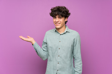 Young man over isolated purple wall holding copyspace imaginary on the palm to insert an ad