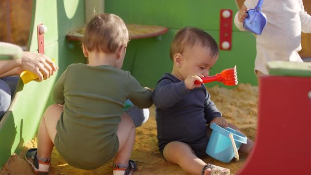 Medium shot of three toddlers in bodysuits and sandals playing together outdoors in sand box, and unrecognizable woman sitting nearby