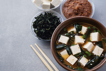 Bowl with miso soup, wakame seaweed, miso pasta, tofu and chopsticks on a gray background.