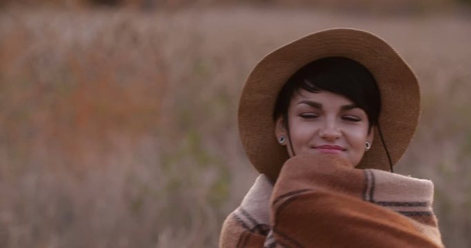Close up portait of attractive short-haired Caucasian hipster girl in straw hat  wraping herself in a warm cozy checkered woolen plaid. It got cooler in the evening. 4K 50 fps slow motion
