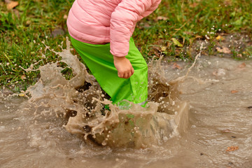Low section of a child girl in green waterproof pants and rubber boots jumping into a massive rain puddle with the water splashing high up in the air on a rainy autumn day in October in Germany