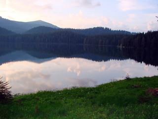 Fototapeta premium A view of the sky with illuminated clouds reflected on the surface of a mountain lake in the middle of the forest at sunset.