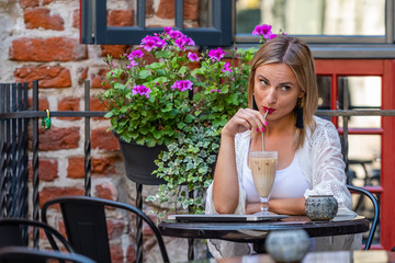 Beautiful blond woman dressed in  light clothes sitting in outdoor cafe and sipping a cocktail.