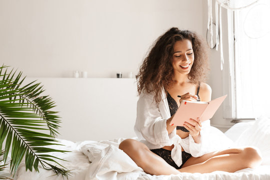 Image of woman wearing lingerie holding diary book on bed in apartment