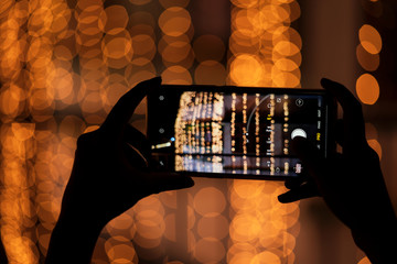 Woman Clicking Photos With Her Smartphone. Raised hand taking wonderful blurred bright yellow orange bokeh background images of festive season Diwali, Christmas, Happy New Year with space for text.