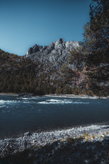 A vertical shot of an amazing autumn landscape in Altai mountains, Russia: rocky shore in the foreground, Katun river in the middle, mountain ridge overgrown with coniferous trees in the background