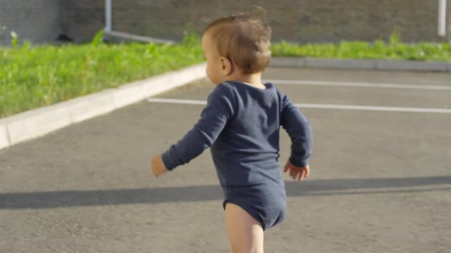 Rear Tracking Shot Of Toddler In Bodysuit Running Around Outdoors On Concrete Lot, And Mom With Twin Sibling Standing Beside