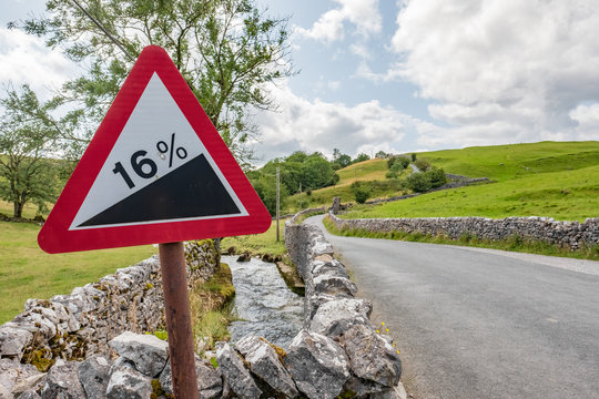 Detailed view of a red warning roadside triangle showing a 16% steep gradient, seen in a rural area of the Yorkshire Dales. Rolling hills can be seen.