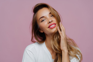 Headshot of carefree relaxed feminine lady tilts head left, touches her long wavy hair, wears...