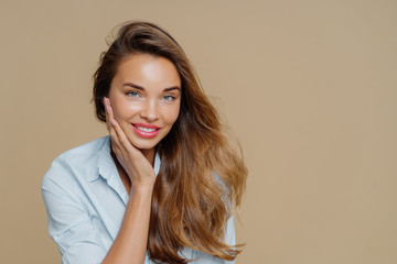 Portrait of cheerful smiling woman touches cheek, has toothy charming smile, wears shirt, looks at camera with pleasure, has long wavy hair, makeup, poses against brown background, free space