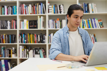 Portrait of young Latin-American man using laptop while studying in college library sitting against bookshelves, copy space