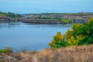 Fototapeta premium Autumn landscape with calm river, cliffs and colorful – green, orange and yellow foliage on the trees. Fall nature.