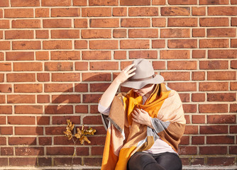 Young woman with a brown hat sitting in front of a brick wall wearing autumn colors looking down